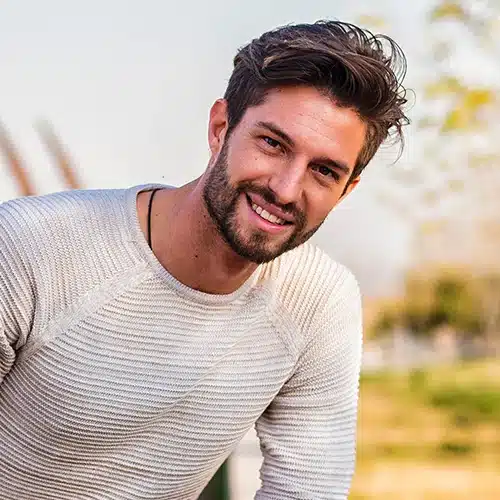 Smiling man with beard wearing a beige ribbed sweater and ripped black jeans posing in a sunlit park.